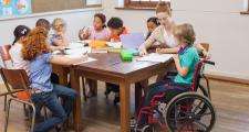 Children sit around a table. A child in a wheelchair is in the front of the frame being helped by a teacher